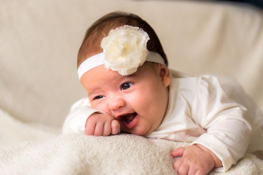 Childhood, motherhood, emotions, fashion concepts - Cute smiling happy chubby baby close up little girl in beautiful white dress flower headband on head lying on soft warm bed on her tummy at home.
