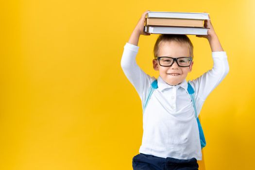 Banner Funny Preschool Child Boy in Glasses with Book on Head and Bag on Yellow Background Copy Space. Happy smiling kid go back to school, kindergarten. Success, motivation, winner, genius concept