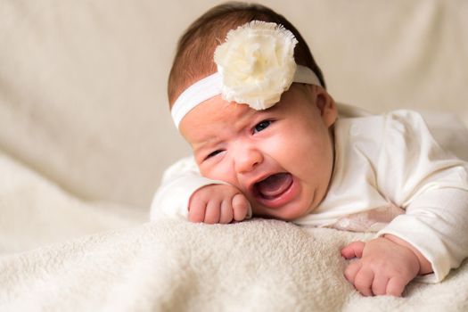 Childhood, motherhood, emotions, fashion concept - worried sad crying chubby baby with open mouth close up little girl in beautiful white dress floral headband lying on soft warm bed on tummy at home.