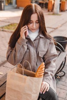 A stylish brunette woman walks around the autumn city. The brunette is sitting on a bench outside with a glass of coffee and a phone, holding a craft bag with a baguette.