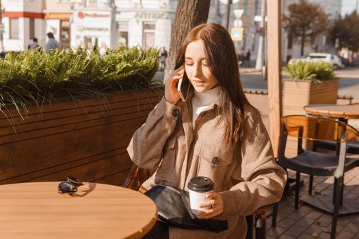 A stylish brunette woman is sitting at a table in an outdoor cafe with coffee and a phone. Autumn walk.