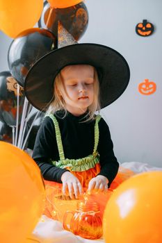 Children's Halloween - a girl in a witch hat and a carnival costume with airy orange and black balloons at home. Ready to celebrate Halloween.