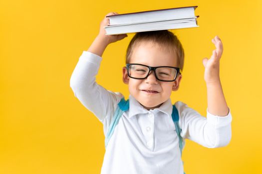 Funny Preschool Child Boy in Glasses with Book on Head and Bag on Yellow Background Copy Space. Happy Smiling Kid Go Back to School, Kindergarten. Success, Motivation, Winner Genius, Superhero concept.