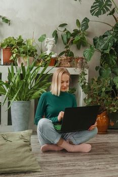 A blonde woman in a room with a lot of green indoor plants is working on a laptop. The concept of biophysical design in the interior. Work from home, work as a freelancer.
