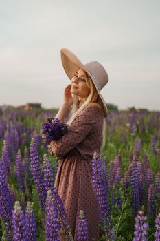 A beautiful woman in a straw hat walks in a field with purple flowers. A walk in nature in the lupin field.