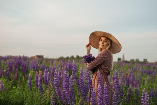 A beautiful woman in a straw hat walks in a field with purple flowers. A walk in nature in the lupin field.