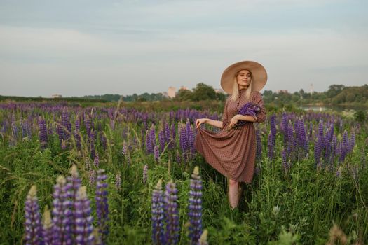 A beautiful woman in a straw hat walks in a field with purple flowers. A walk in nature in the lupin field.