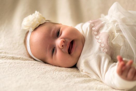 Childhood, motherhood, emotions, fashion concept - worried sad crying chubby baby with open mouth close up little girl in beautiful white dress floral headband lying on soft warm bed on tummy at home.