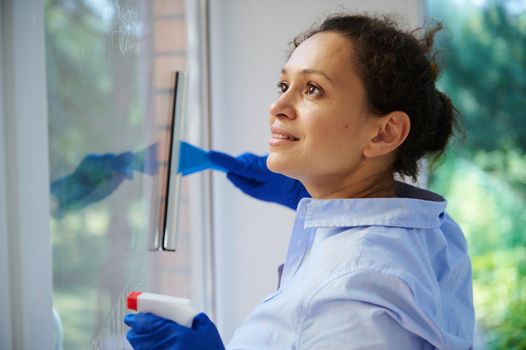 Close-up of multi-ethnic pretty woman, maid, housewife, homemaker spraying detergent and removing stains while washing windows in the veranda. Housekeeping. Household. Chores. Spring cleaning concept