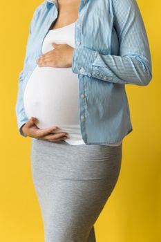 Motherhood, femininity, love, care, waiting, hot summer - bright croped Close-up unrecognizable pregnant woman in shirt with small baby shoes hand over tummy rub belly on yellow background, copy space.