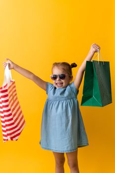 Portrait Beautiful Happy Little Preschool Girl In Sunglasses Smiling Cheerful Holding Lift Up Cardboard Bags Isolated On Orange Yellow Background. Happiness, Consumerism, Sale People shopping Concept.