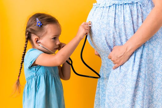 Motherhood, hot summer - croped portrait pregnant mother unrecognizable woman blue dress little Daughter girl sibling treat mom role play game stethoscope unborn big belly tummy on yellow background.