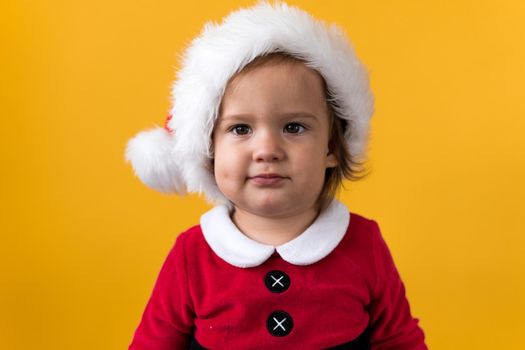 Smiling Portraite Cute Happy Cheerful Chubby Baby Girl in Santa Hat Looking On Camera At Yellow Background. Child Play Christmas Scene Celebrating Birthday. Kid Have Fun Spend New Year Time Copy Space.