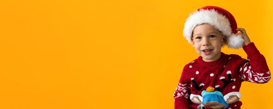 Portrait of happy smiling positive joyful positive preschool little boy in red warm santa hat showing thumb up on orange, yellow background. Winter, holiday, celebration, Christmas, New Year copy space.