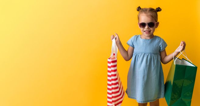 Portrait Beautiful Happy Little Preschool Girl In Sunglasses Smiling Cheerful Holding Cardboard Bags Isolated On Orange Yellow Studio background. Happiness, Consumerism, Sale People shopping Concept.