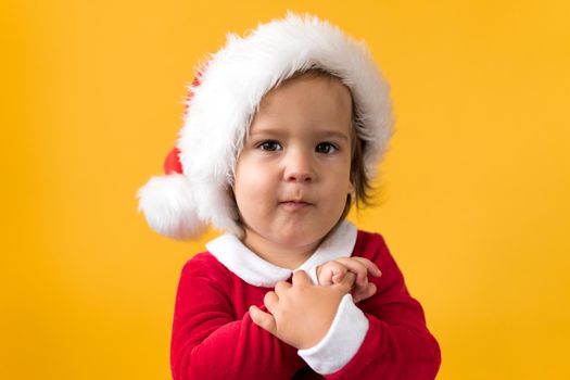 Smiling Portraite Cute Happy Cheerful Chubby Baby Girl in Santa Hat Looking On Camera At Yellow Background. Child Play Christmas Scene Celebrating Birthday. Kid Have Fun Spend New Year Time Copy Space.