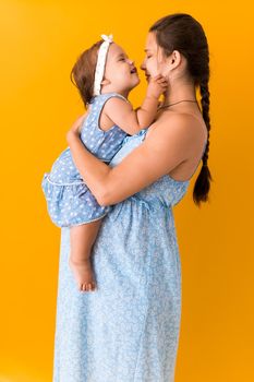 Motherhood, hot summer - portrait young pregnant happy smiling joy mother woman in blue dress holding little baby daughter toddler sibling bare feet in arms looking into eyes kiss on yellow background.