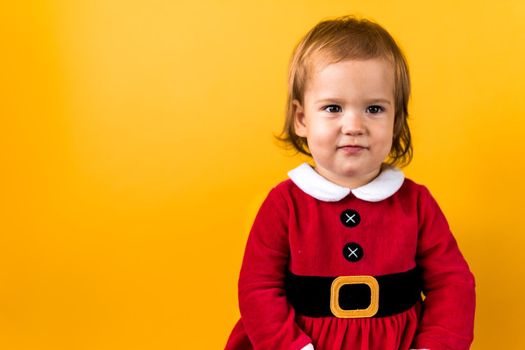 Banner Portraite Cute Happy Cheerful Chubby Baby Girl in Santa Suit Looking On Camera At Yellow Background. Child Play Christmas Scene Celebrating Birthday. Kid Have Fun Spend New Year Time Copy Space.