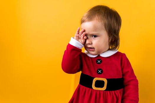 Banner Portraite Cute Happy Cheerful Chubby Baby Girl in Santa Suit Sleepy Rubs Eyes At Yellow Background. Child Play Christmas Scene Celebrating Birthday. Kid Have Fun Spend New Year Time Copy Space.