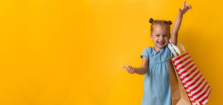 Banner Portrait Beautiful Happy Little Preschool Girl Smiling Cheerful Holding Cardboard Bags Points Finger To Side Up On Orange Yellow Background. Happiness, Consumerism, Sale People shopping Concept.