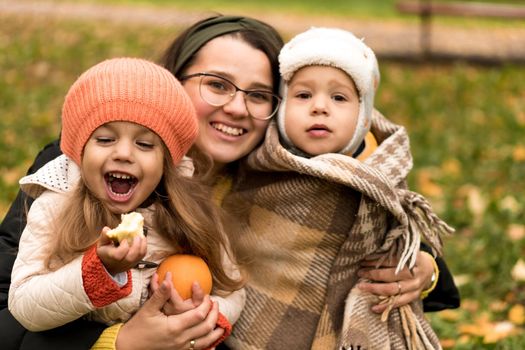 Young Woman Mom And Little Cute Preschool Minor Children In Orange Plaid At Yellow Fallen Leaves Nice Smiling Look At Camera In Cold Weather In Fall Park. Childhood, Family, Motherhood, Autumn Concept.