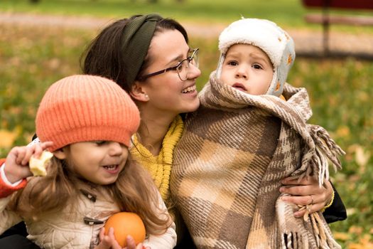 Young Woman Mom And Little Cute Preschool Minor Children In Orange Plaid At Yellow Fallen Leaves Nice Smiling Look At Camera In Cold Weather In Fall Park. Childhood, Family, Motherhood, Autumn Concept.