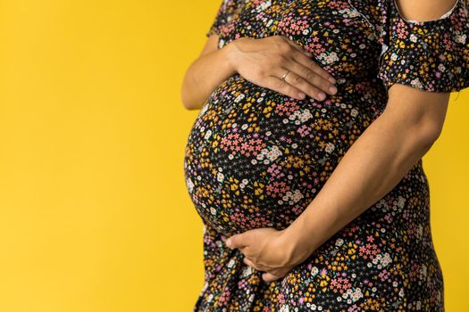 Motherhood, femininity, love, care, waiting, hot summer - bright croped Close-up unrecognizable pregnant woman in floral black dress with hands over tummy rub belly on yellow background, copy space.