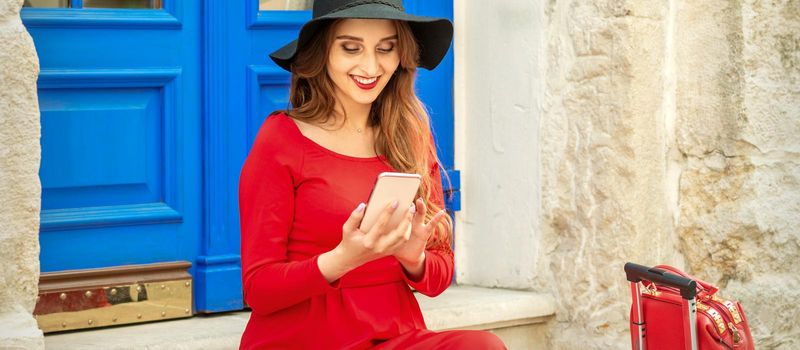 Beautiful young caucasian traveling woman in black hat looking on the smartphone smiling and sitting on stairs at the door outdoors