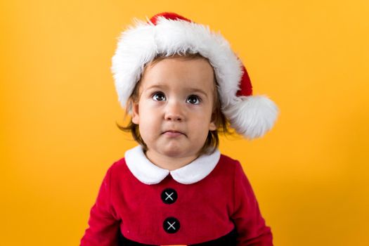 Smiling Portraite Cute Cheerful Chubby Baby Girl in Santa Hat Looking Up Interested At Yellow Background. Child Play Christmas Scene Celebrating Birthday. Kid Have Fun Spend New Year Time Copy Space.