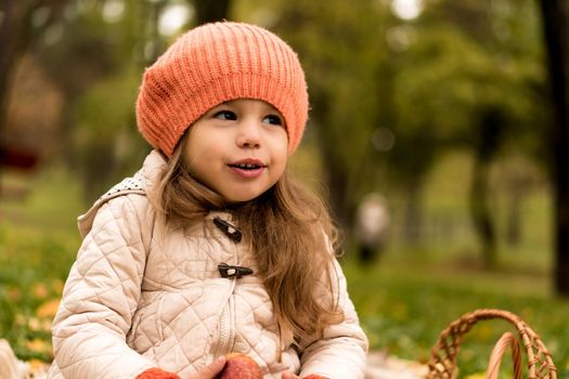 Close Up Portrait Little Cute Preschool Minor Girl In Orange Beret On Yellow Fallen Leaves Eats Red Apple Dreamily Looks Away. Cold Weather In Fall Park. Childhood, Family, Motherhood, Autumn Concept.