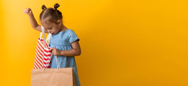 Banner Portrait Caucasian Beautiful Happy Little Preschool Girl Smiling Cheerful And Holding Cardboard Bags Isolated On Orange Yellow Background. Happiness, Consumerism, Sale People shopping Concept.