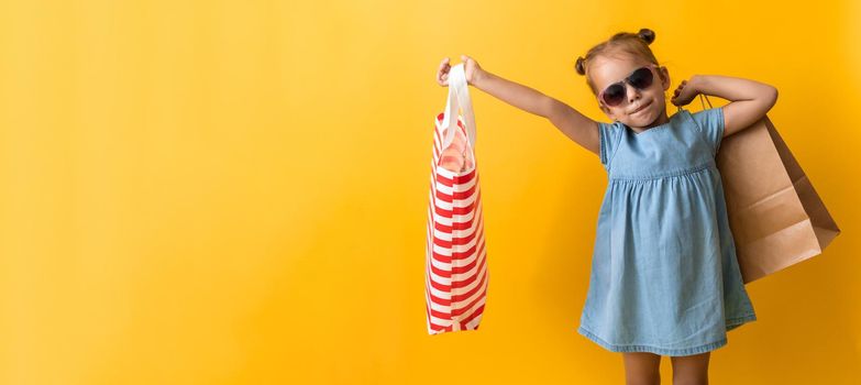 Portrait Beautiful Happy Little Preschool Girl In Sunglasses Smiling Cheerful Holding Cardboard Bags Isolated On Orange Yellow Studio background. Happiness, Consumerism, Sale People shopping Concept.