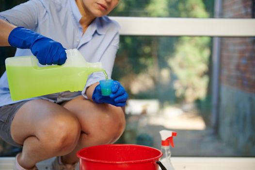 Details: hands of a housewife, homemaker, maid pouring cleaner detergent into a red bucket, for washing and mopping the floor while cleaning the house. Housekeeping. Chores. Household. Housework.