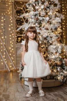 A girl in a festive light dress with a gift in her hands next to a Christmas tree, lights of garlands in the background. The concept of New Year holidays.