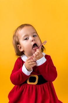 Portraite Cute Cheerful Chubby Baby Girl in Santa Suite Holding Eating Caramel Candy At Yellow Background. Child Play Christmas Scene Celebrating Birthday. Kid Have Fun Spend New Year Time Copy Space.