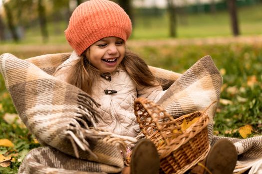 Little Cute Preschool Minor Girl In Orange Beret On Plaid At Yellow Fallen Leaves Basket Laughs Nice Smiling Look At Camera In Cold Weather In Fall Park. Childhood, Family, Motherhood, Autumn Concept.