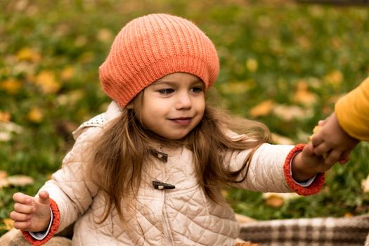 Portrait Of Little Cute Preschool Minor Girl In Orange Beret On Yellow Fallen Leaves Takes Apple From Someone Looks Away At Cold Weather In Fall Park. Childhood, Family, Motherhood, Autumn Concept.