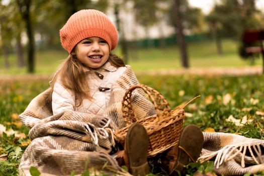 Little Cute Preschool Minor Girl In Orange Beret On Plaid At Yellow Fallen Leaves Basket Laughs Nice Smiling Look At Camera In Cold Weather In Fall Park. Childhood, Family, Motherhood, Autumn Concept.