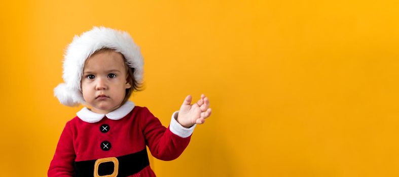 Banner Serious Portraite Cute Fun Cheerful Chubby Baby Girl in Santa Hat Waving Hand At Yellow Background. Child Play Christmas Scene Celebrating Birthday. Kid Have Fun Spend New Year Time Copy Space.