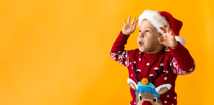 Portrait of happy smiling positive joyful positive preschool little boy in red warm santa hat showing thumb up on orange, yellow background. Winter, holiday, celebration, Christmas, New Year copy space.