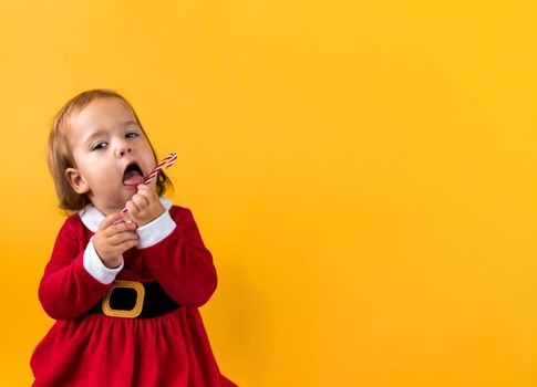 Banner Portraite Cute Cheerful Chubby Baby Girl in Santa Suite Holding Eating Caramel Candy At Yellow Background. Child Play Christmas Celebrating Birthday. Kid Have Fun Spend New Year Time Copy Space.