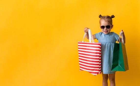 Portrait Beautiful Happy Little Preschool Girl In Sunglasses Smiling Cheerful Holding Cardboard Bags Isolated On Orange Yellow Studio background. Happiness, Consumerism, Sale People shopping Concept.