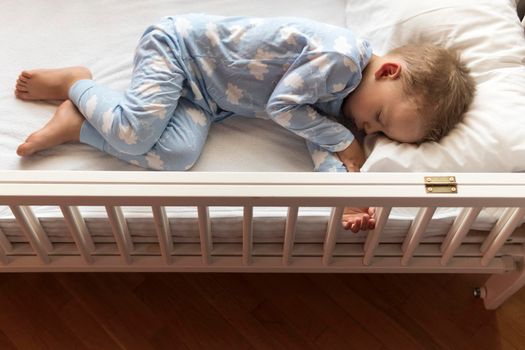 top view Cute little 2-3 years preschool baby boyl kid sleeping sweetly in white crib during lunch rest time in blue pajama with pillow at home. Childhood, leisure, comfort, medicine, health concept.