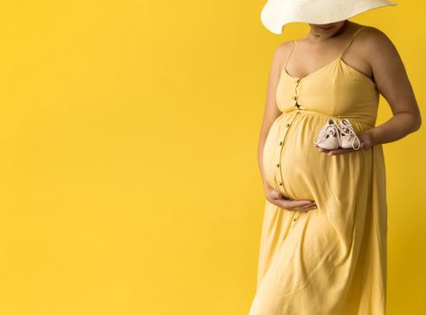 Motherhood, femininity, fashion,, hot summer - banner Close-up unrecognizable pregnant woman lady in beautiful vintage dress Provence straw hat with small baby shoes on yellow background, copy space.