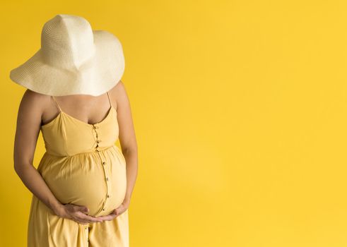 Motherhood, femininity, fashion, hot summer - banner unrecognizable pregnant woman lady in beautiful vintage dress Provence straw hat with hands over tummy rubs tummy on yellow background copy space.