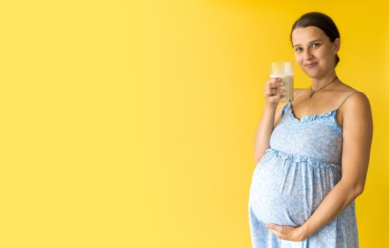 portrait cute beautiful pregnant young pretty woman in floral blue dress holds drink glass of milk rubs tummy on yellow background. Motherhood, femininity, healthy eating food, dairy, summer concept.