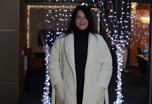 Cheerful brunette plus-size indoors in black sweater and white winter coat has her hands in her pockets and looks at the camera.