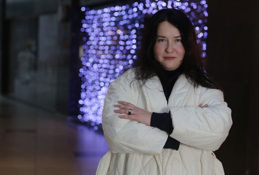 Attractive brunette plus size indoors wearing a black sweater and white winter coat looking at the camera with her arms crossed. Portrait of a woman in a white coat in a store.