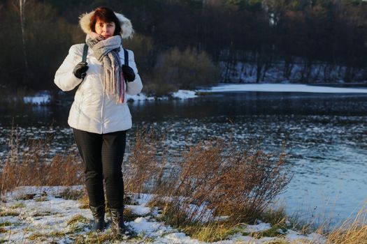 Woman in white jacket with scarf and black gloves standing near river in forest.