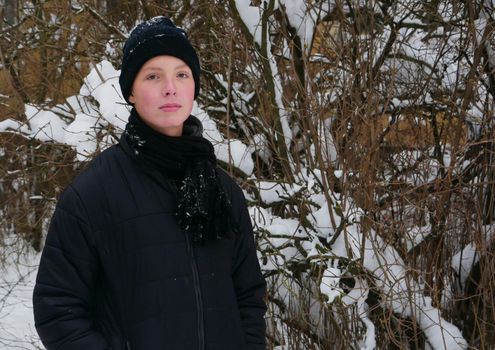 Portrait of a boy in a black winter sports jacket, scarf, hat against the background of tree branches in the snow.